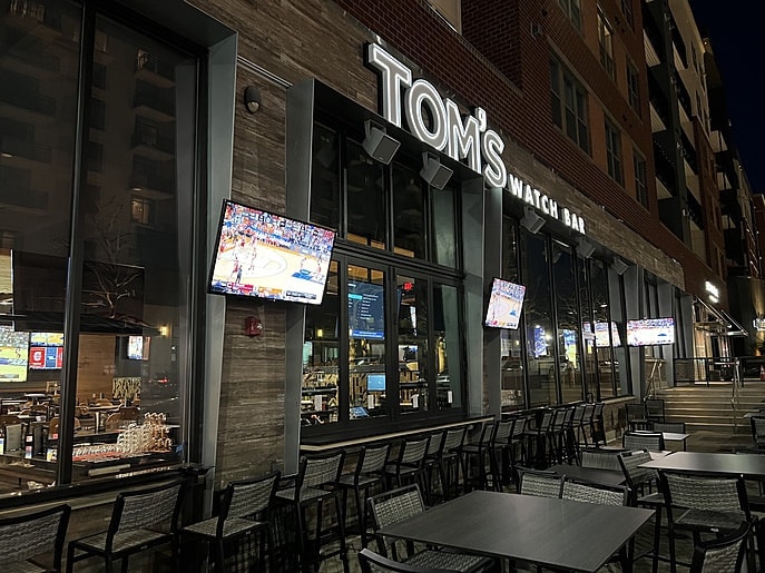 Exterior of a restaurant with a neon sign that reads “Tom’s Watch Bar” and outdoor TVs.