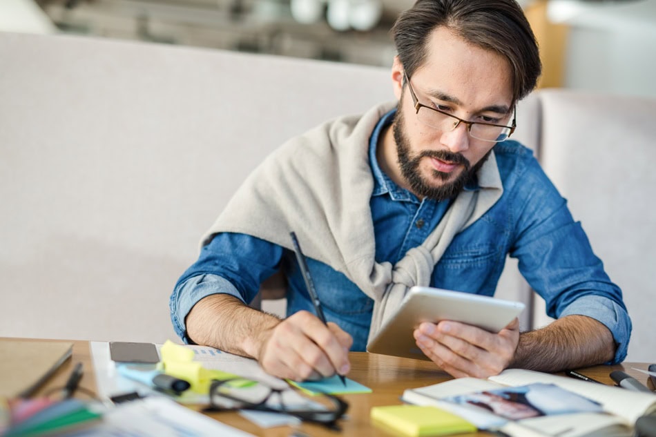 Man writing paper while reading tablet indoors.