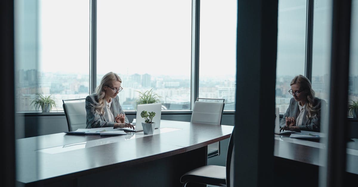 A businesswoman in a blazer works at a desk in a modern office with city views.